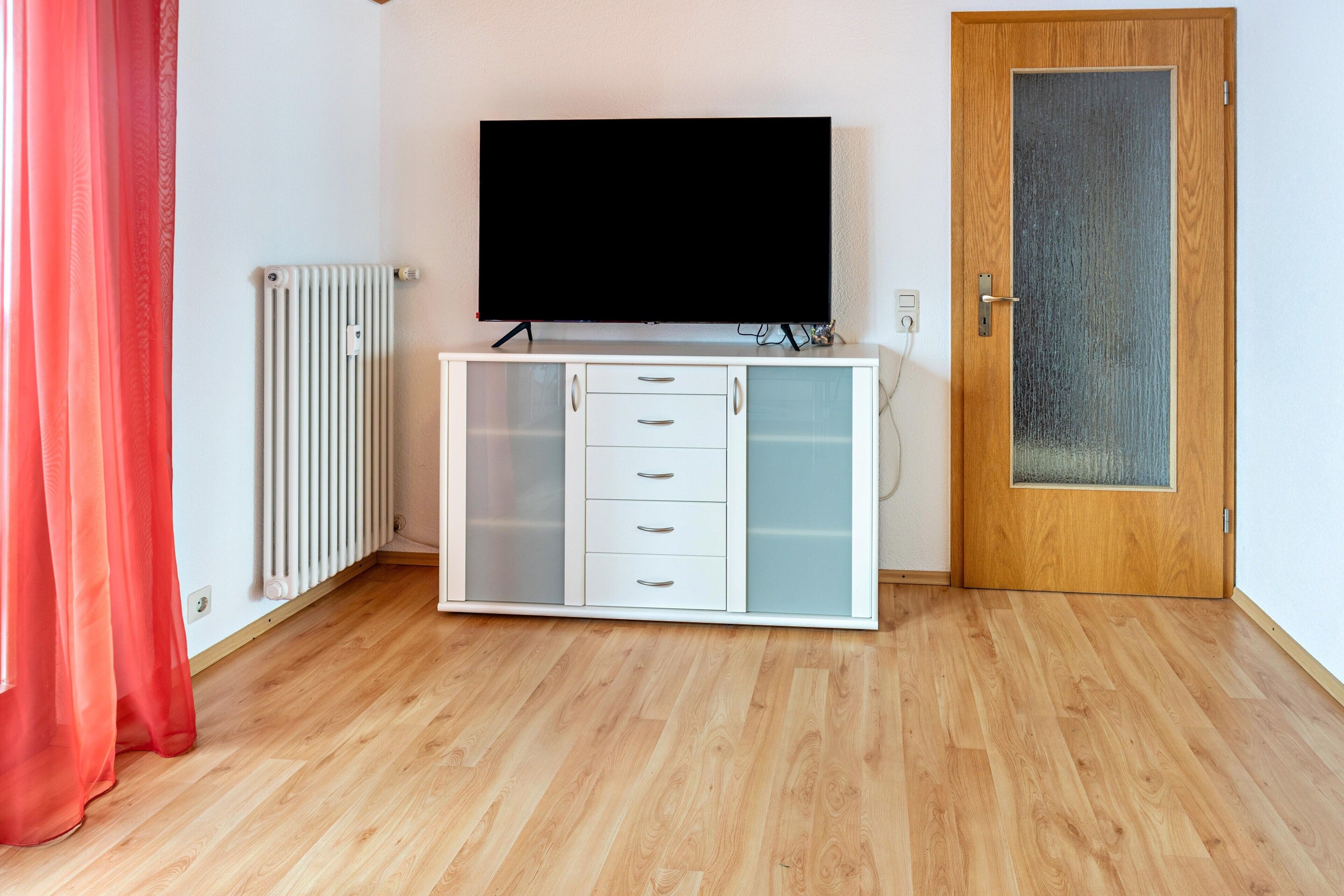 Una habitación con un armario blanco, televisor, puerta de madera y radiador.