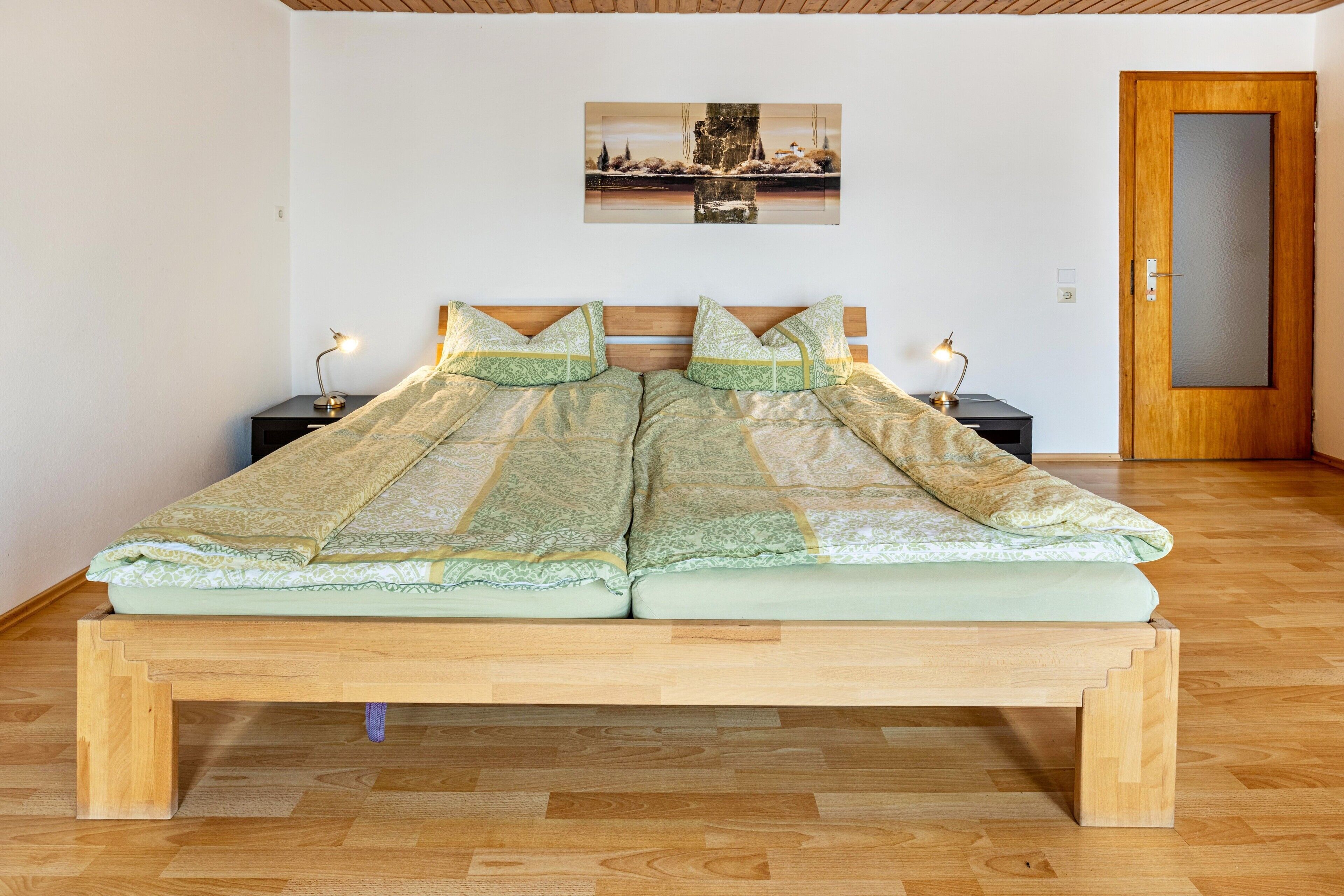 A double bed with a patterned bedspread, two bedside tables with lamps, a wooden floor, and a door.