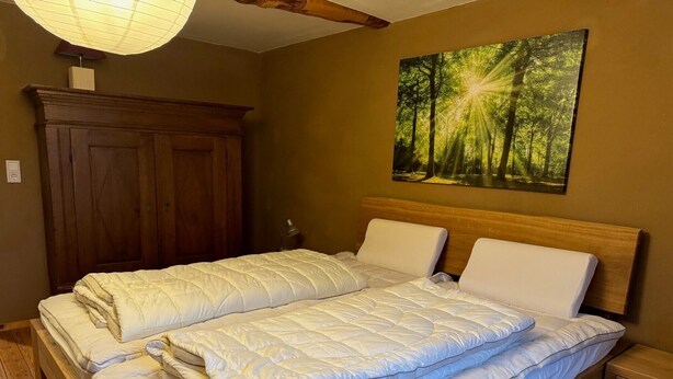 Un lit double avec une couette matelassée, une tête de lit en bois, une table de chevet, une lampe et un tableau représentant une forêt.