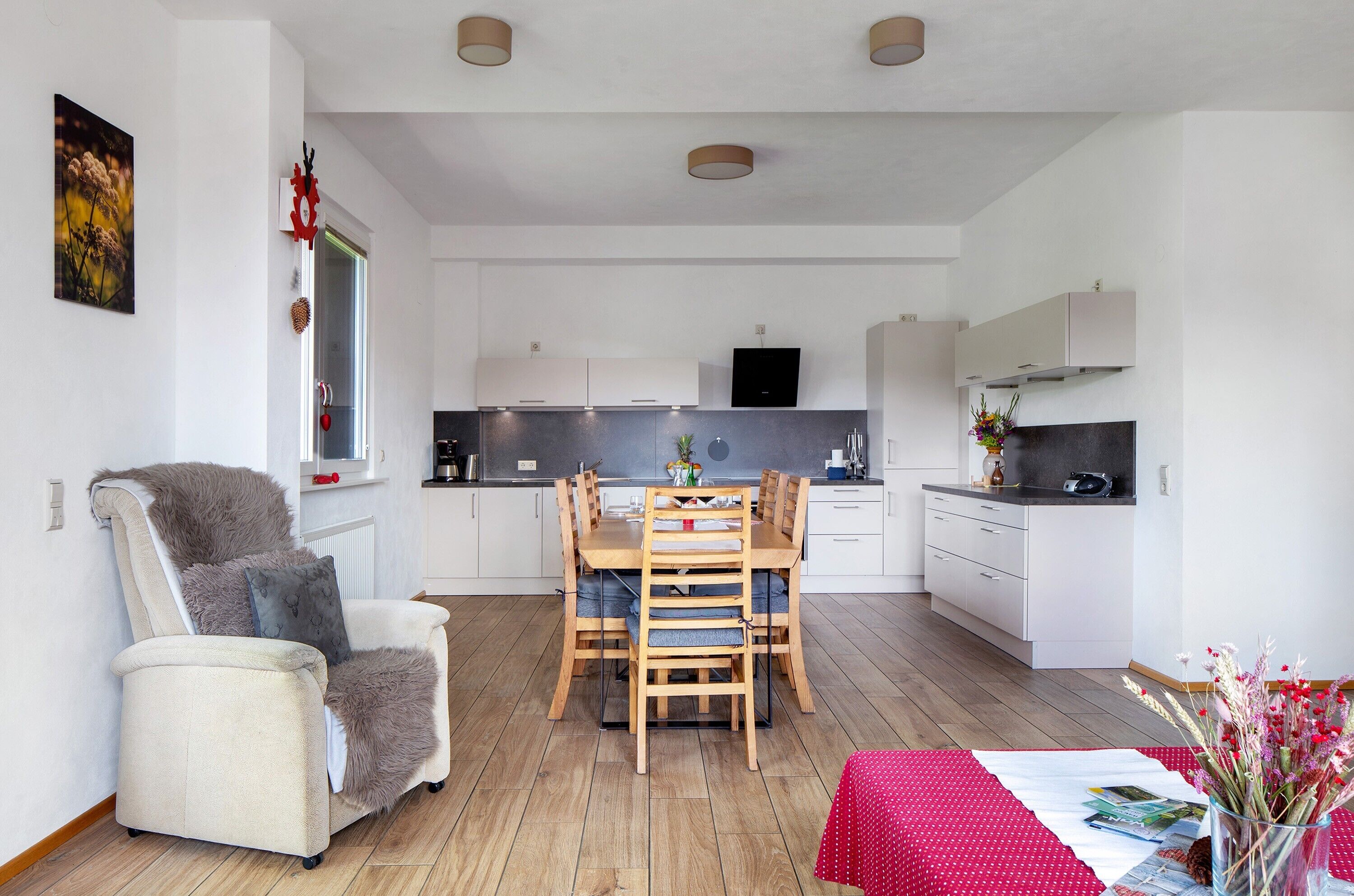 A modern kitchen with white cabinets, a dining table with chairs, a sofa, and a red tablecloth.