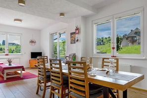 A dining room with a wooden table and chairs, a television, and large windows with a view of a house and greenery. - Apartment 'Ulrich' with Mountain View, Shared Terrace and Wi-Fi (Oberried-Hofsgrund)