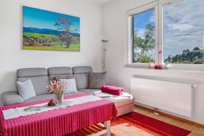 A room with a sofa, a table with a red tablecloth, a window with a view of a landscape, and a radiator. - Apartment 'Ulrich' with Mountain View, Shared Terrace and Wi-Fi (Oberried-Hofsgrund)