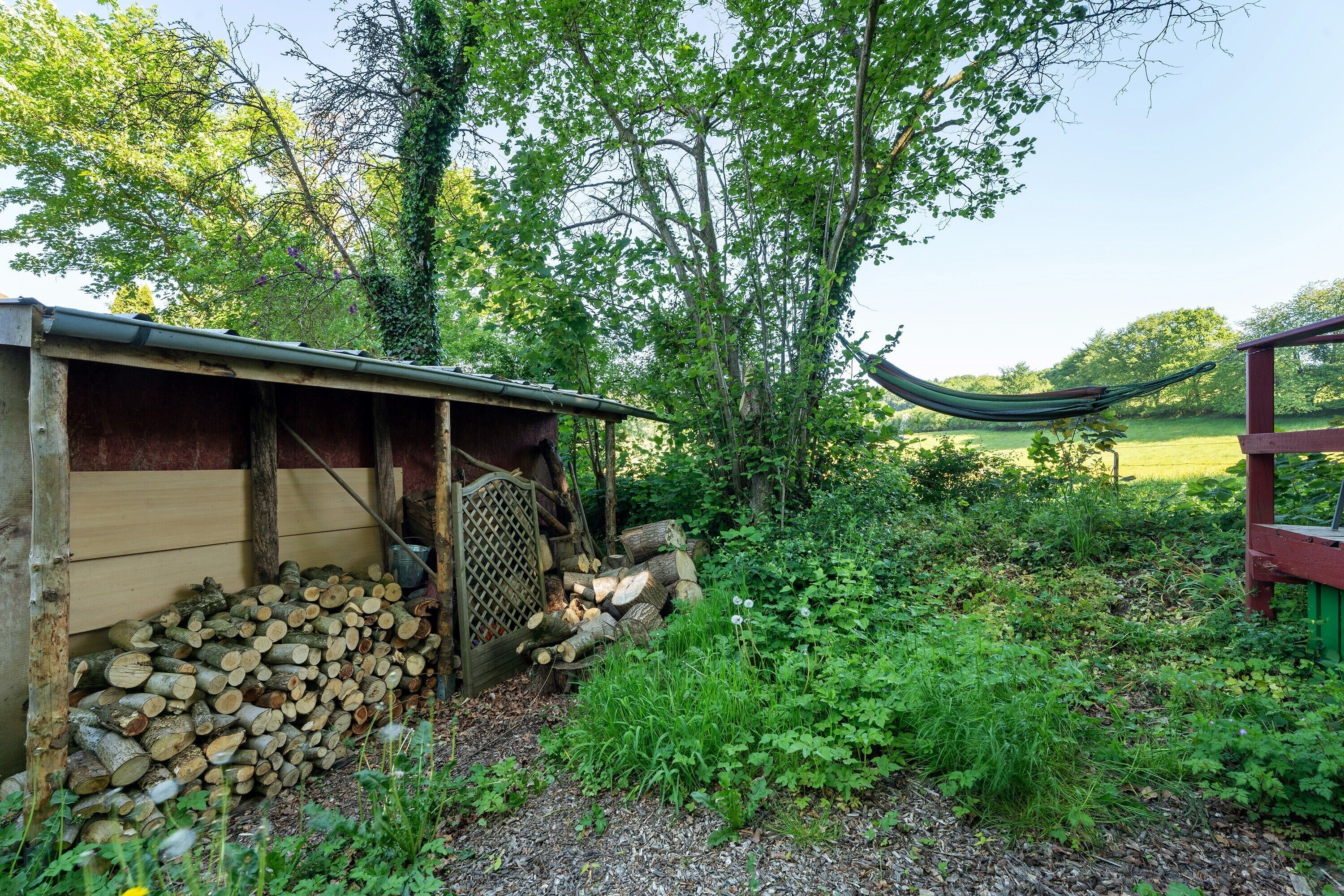 A rustic wooden structure with a pile of logs, a hammock, and a garden area with various plants.