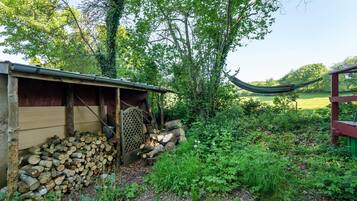 A rustic wooden structure with a pile of logs, a hammock, and a garden area with various plants.