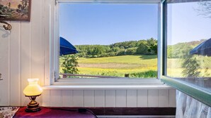 Une fenêtre donnant sur un paysage verdoyant, une lampe sur une table et un tableau encadré au mur.
