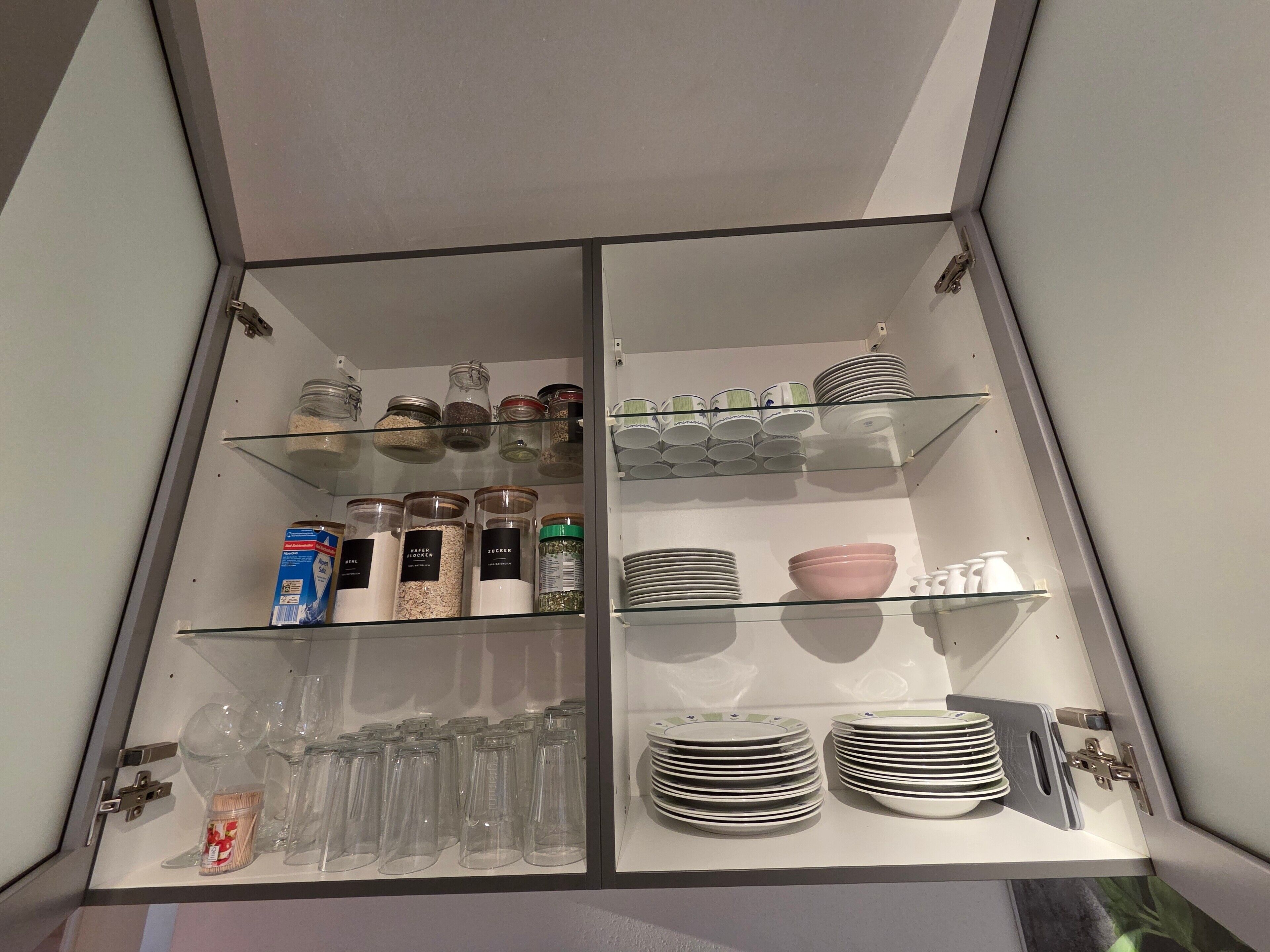 Un armario de cocina con repisas de vidrio que contiene frascos, platos y utensilios.