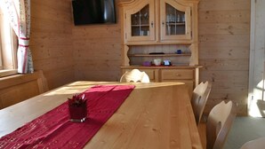 A wooden cabin interior with a dining table, chairs, a television mounted on the wall, and a wooden cabinet with glass doors.