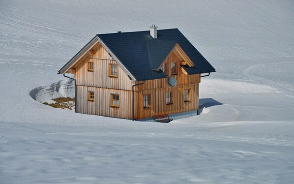 A wooden cabin with a steeply pitched roof, multiple windows, and a chimney, situated on a snow-covered slope.