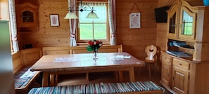 A wooden cabin interior with a dining table, benches, a window with curtains, a wall-mounted lamp, and a cabinet with a guitar.