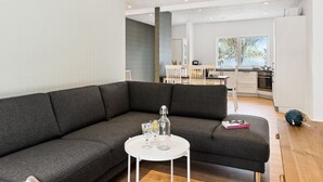 En moderne stue med en grå hjørnesofa, et lille hvidt bord med glas og en flaske, og et køkken i baggrunden med et vindue der viser et sneklædt landskab.