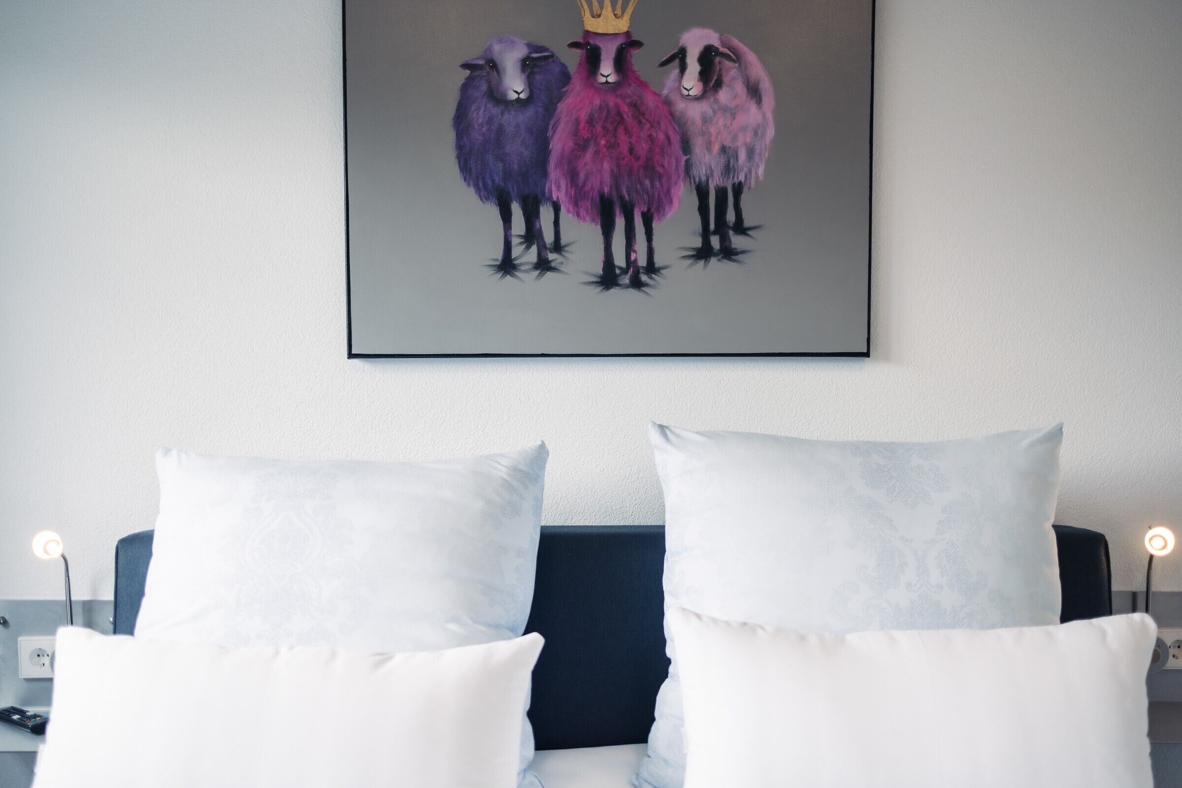 Ein Hotelzimmer mit einem Bett, zwei weißen Kissen, einem dunklen Kopfteil und einem Wandbild mit Schafen.