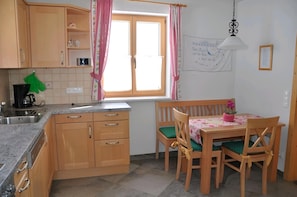 Une cuisine avec des meubles de rangement en bois, un évier, une fenêtre avec des rideaux roses, une table à manger avec des chaises et un luminaire suspendu.