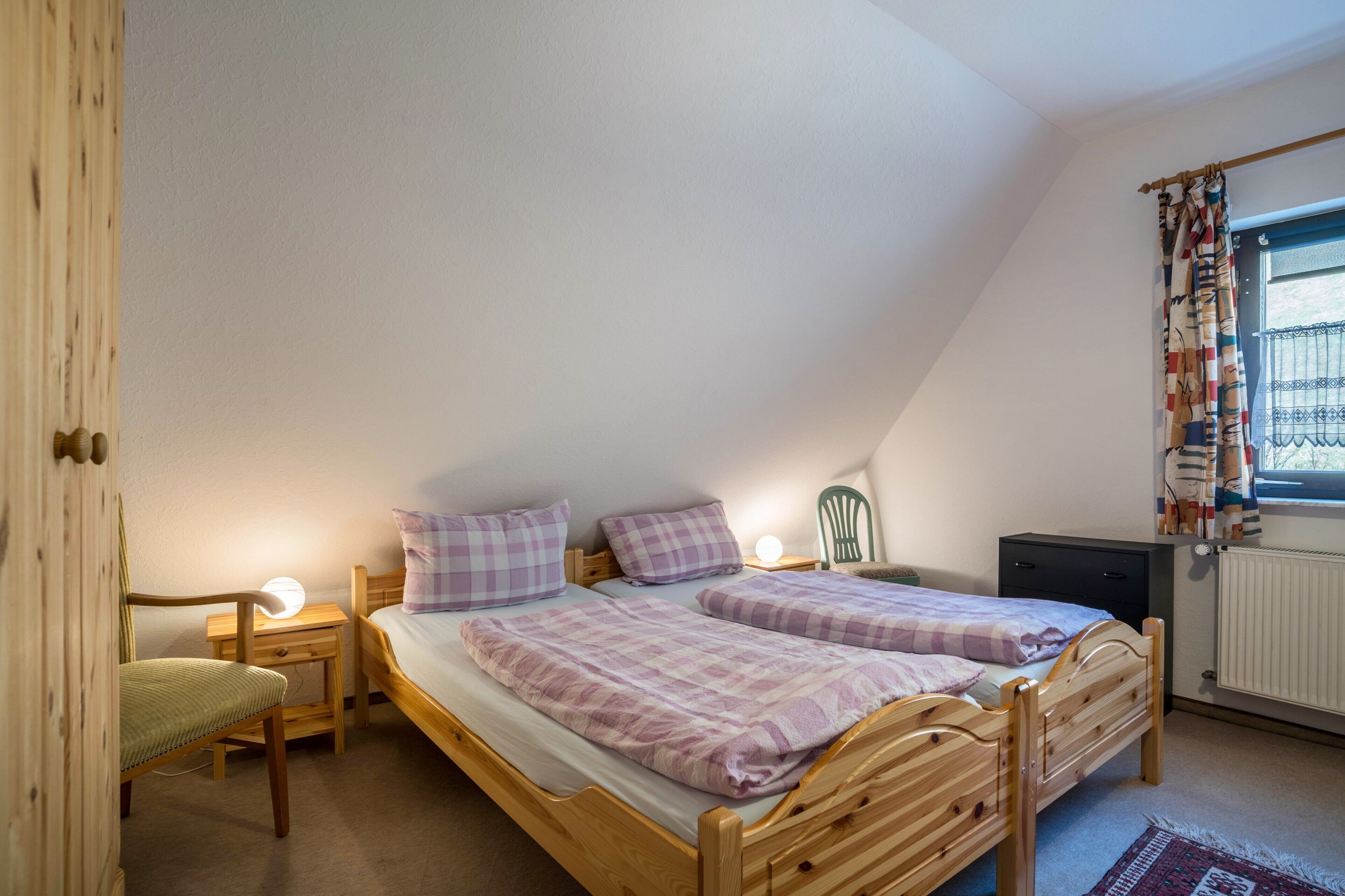 Quarto com duas camas de madeira, uma cadeira, uma mesinha de cabeceira com abajur e uma janela com cortinas.
