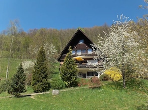 A two-story chalet with a balcony, surrounded by a lush garden and a forested backdrop.