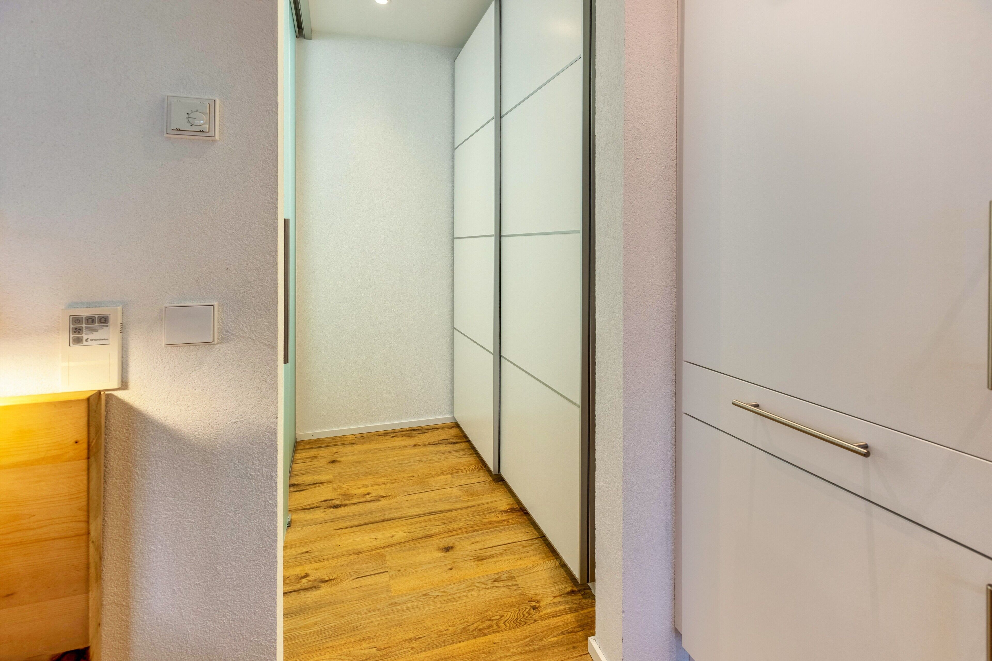 A narrow hallway with wooden flooring, a white wall, and a white cabinet with a gold handle.