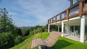 Ein modernes Haus mit einer Holzterrasse, einer überdachten Veranda mit Gartenmöbeln und einem üppigen, grünen Garten.