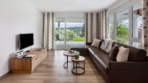 Ein modernes Wohnzimmer mit Flachbildfernseher, Couchtisch aus Holz, Ledersofa und großen Fenstern mit Blick auf eine grüne Landschaft.
