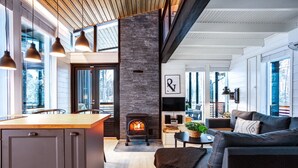 A modern interior with a kitchen, dining area, and living room, featuring a wood-burning stove, large windows, and a cozy seating area.