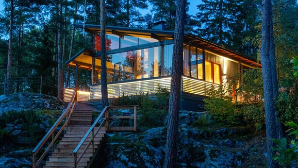 A modern wooden cabin with a glass extension, surrounded by trees and rocks.