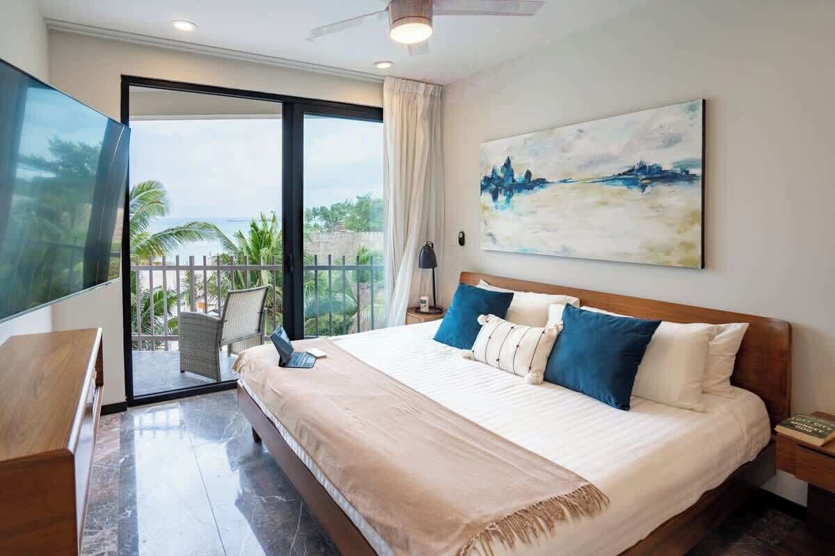 A hotel room with a large bed, a TV, a balcony with a view of palm trees and the sea, and a painting on the wall.