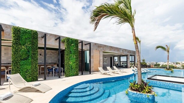 A modern pool area with a curved edge, lounge chairs, and a view of the city. - Chic Studio by 5th Ave with Rooftop Pool and Gym (Playa del Carmen)