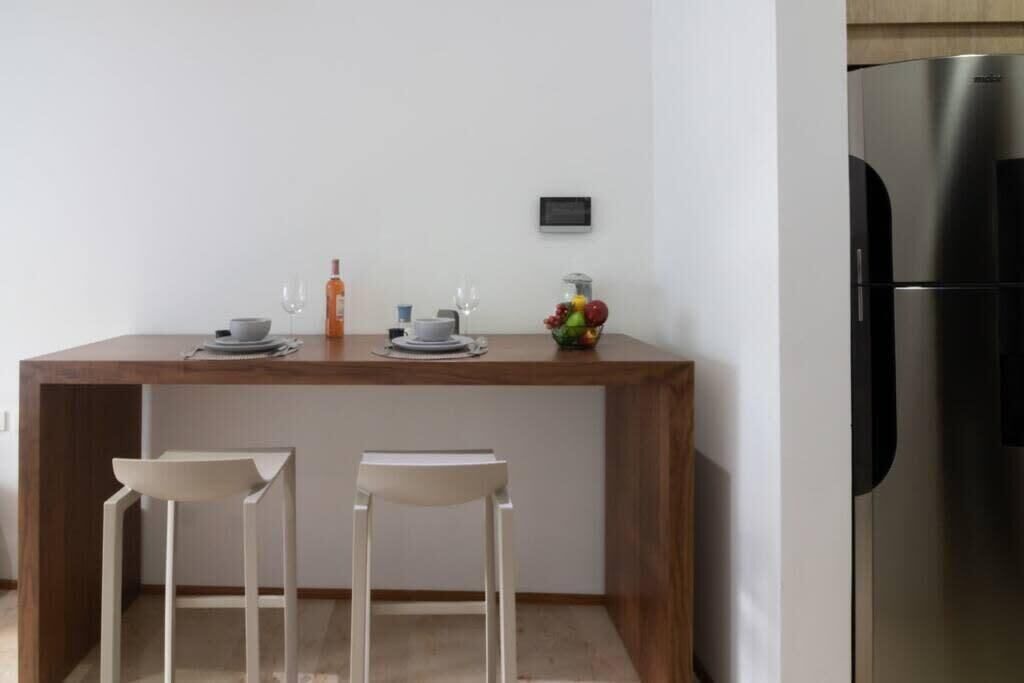 A small kitchen with a wooden countertop, two white stools, a stainless steel refrigerator, and a fruit bowl.