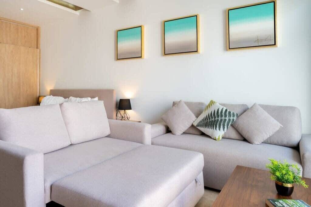 Een moderne woonkamer met een grote L-vormige bank, een klein bijzettafeltje met een plant, en drie ingelijste kunstwerken aan de muur.