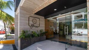 A modern building entrance with a logo, glass doors, and a planter with plants. - Chic Studio by 5th Ave with Rooftop Pool and Gym (Playa del Carmen)