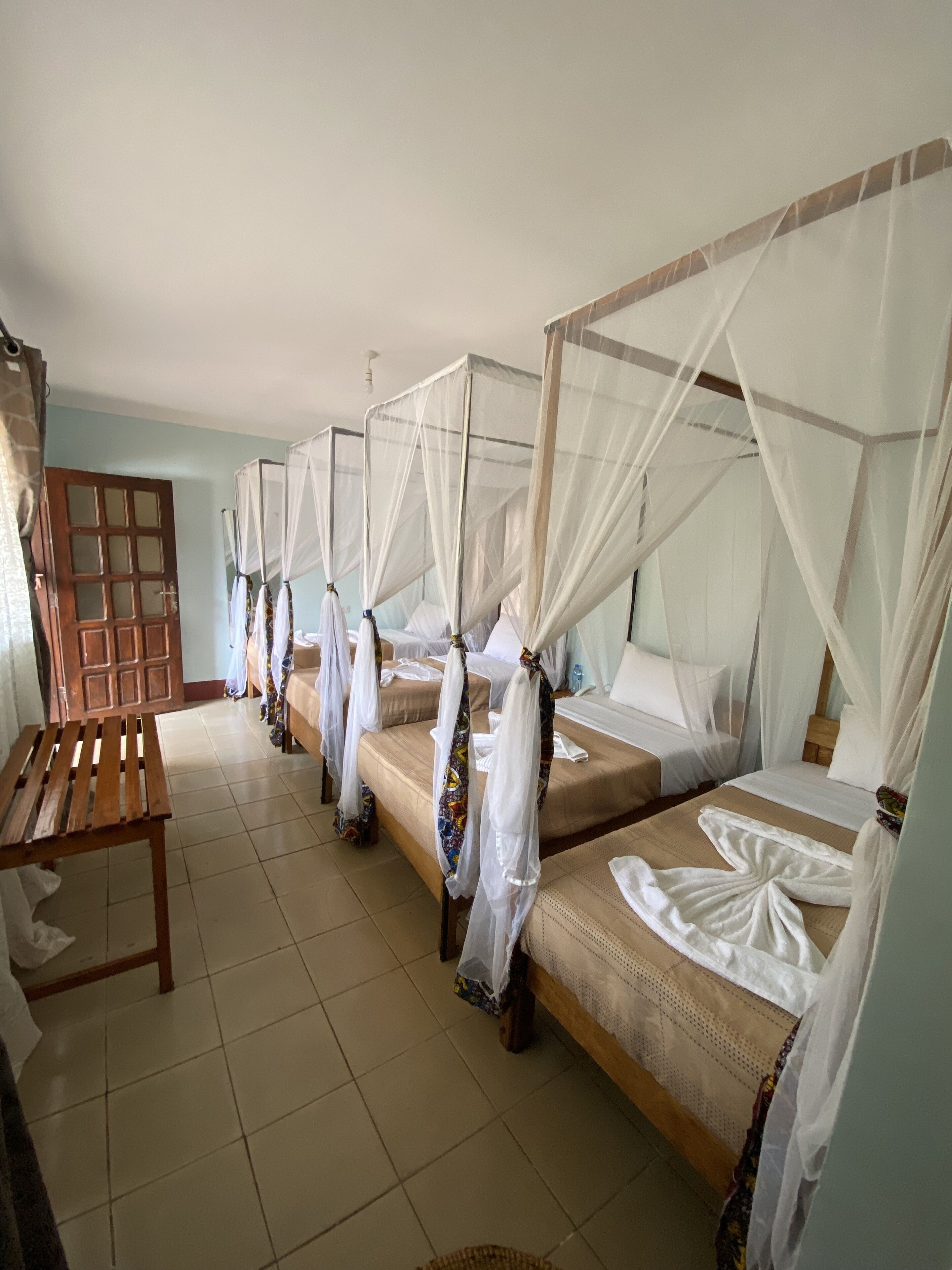 A room with four beds, each with a canopy, a wooden bench, and a tiled floor.