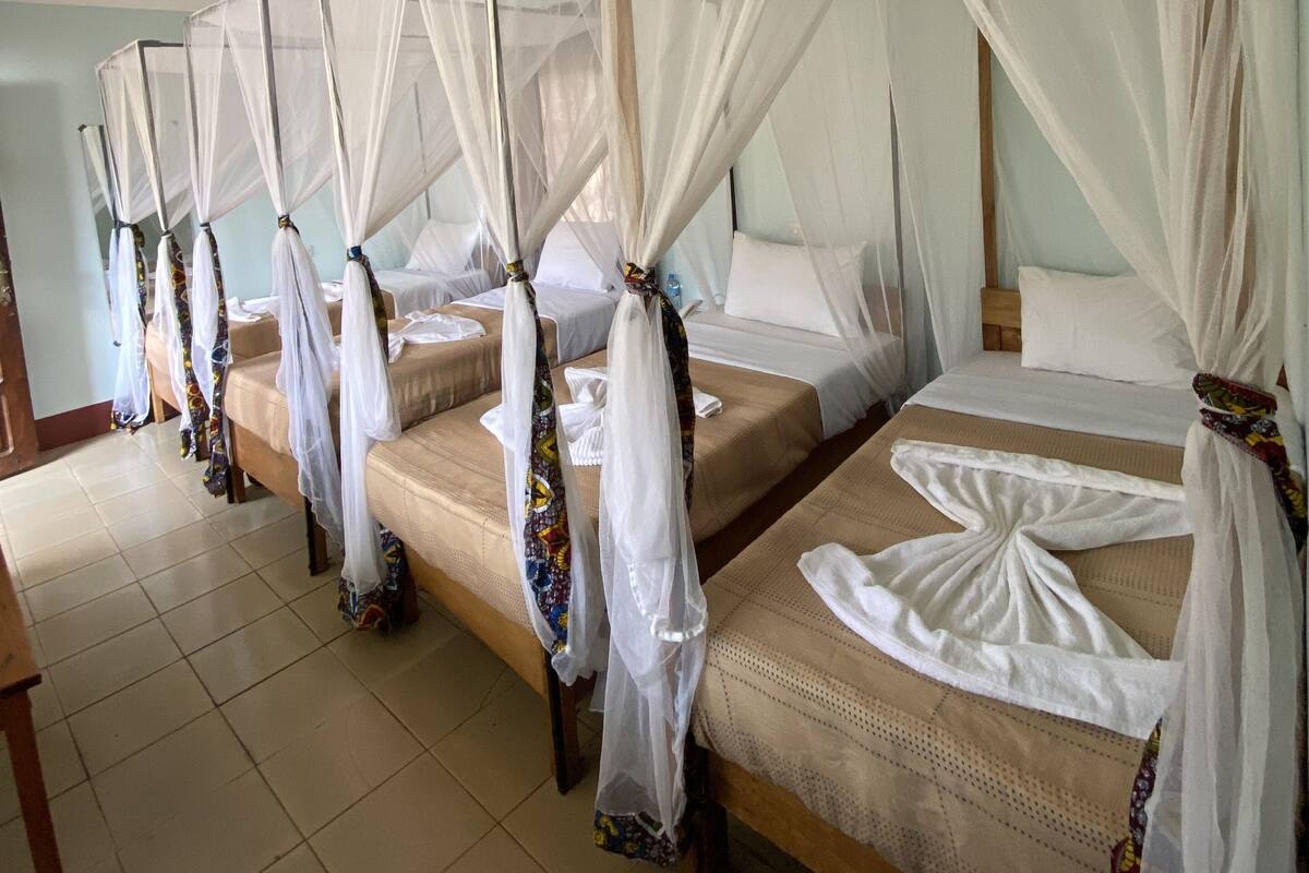 A row of four single beds with white bedsheets and mosquito netting, wooden headboards, and a tiled floor.