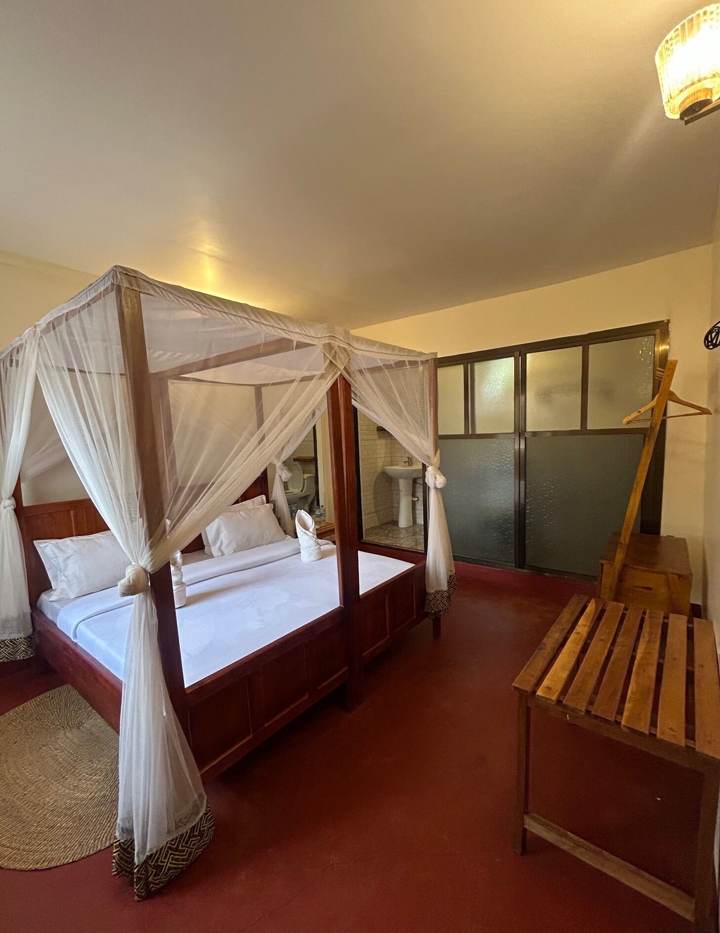 Una cama doble con dosel, un banco de madera y un baño visible a través de una puerta de vidrio.
