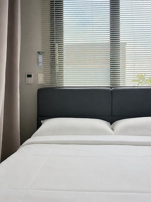 A bed with a dark headboard, a window with blinds, and a thermostat on the wall. - Somsom House (Seogwipo)