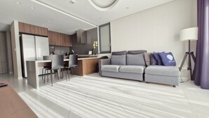 Ein modernes Wohnzimmer mit grauem Sofa, einem Essbereich mit weißem Tisch und Stühlen sowie einer Küche mit dunklen Fliesen und Holzschränken.