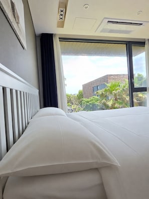 A hotel room with a bed, a window with a view of buildings and greenery, and an air conditioning unit on the ceiling. - Somsom House (Seogwipo)