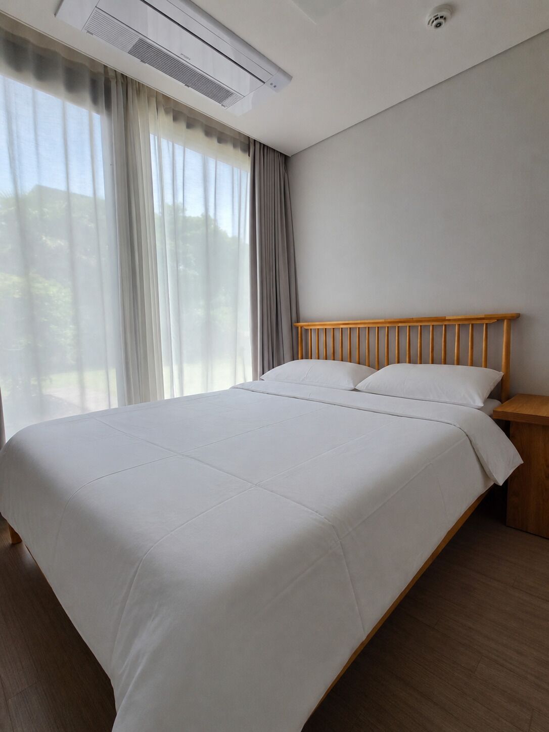 A neatly made bed with a wooden headboard, a bedside table, and a window with curtains.