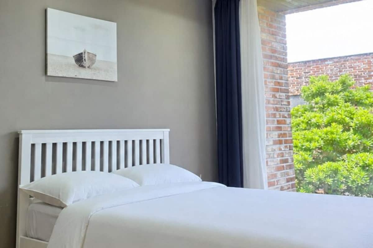 Une chambre dâhĂŽtel moderne avec un lit blanc, un tableau au mur et une vue sur un bĂątiment en briques et sur la verdure environnante.