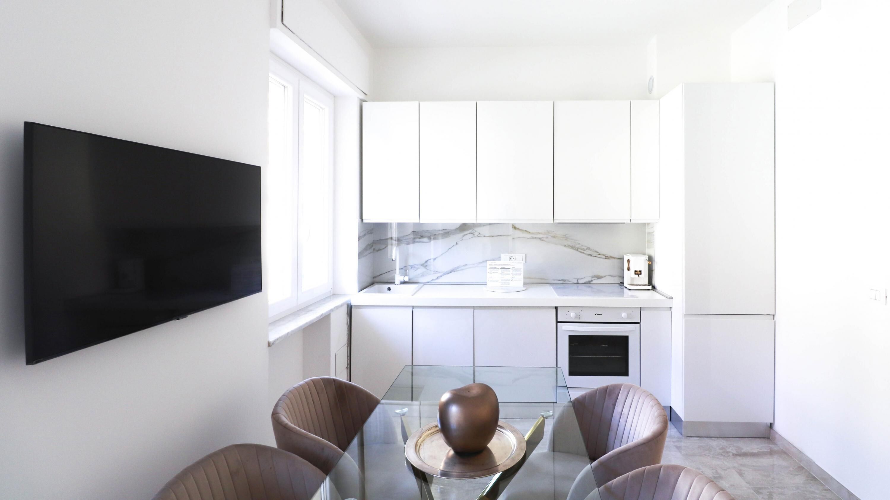 A modern kitchen with white cabinets, a marble backsplash, a built-in oven, and a flat-screen TV mounted on the wall.