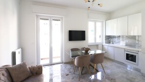 Cocina moderna con armarios blancos, encimera de mármol y horno empotrado. Cuenta con una zona de comedor con mesa de cristal y sillas, y un televisor de pantalla plana montado en la pared.