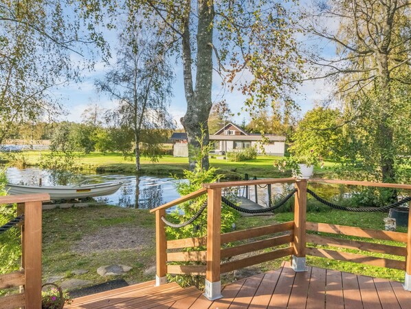 Une terrasse en bois avec une barrière faite de cordes, un bateau sur une étendue d’eau calme et une maison en arrière-plan.