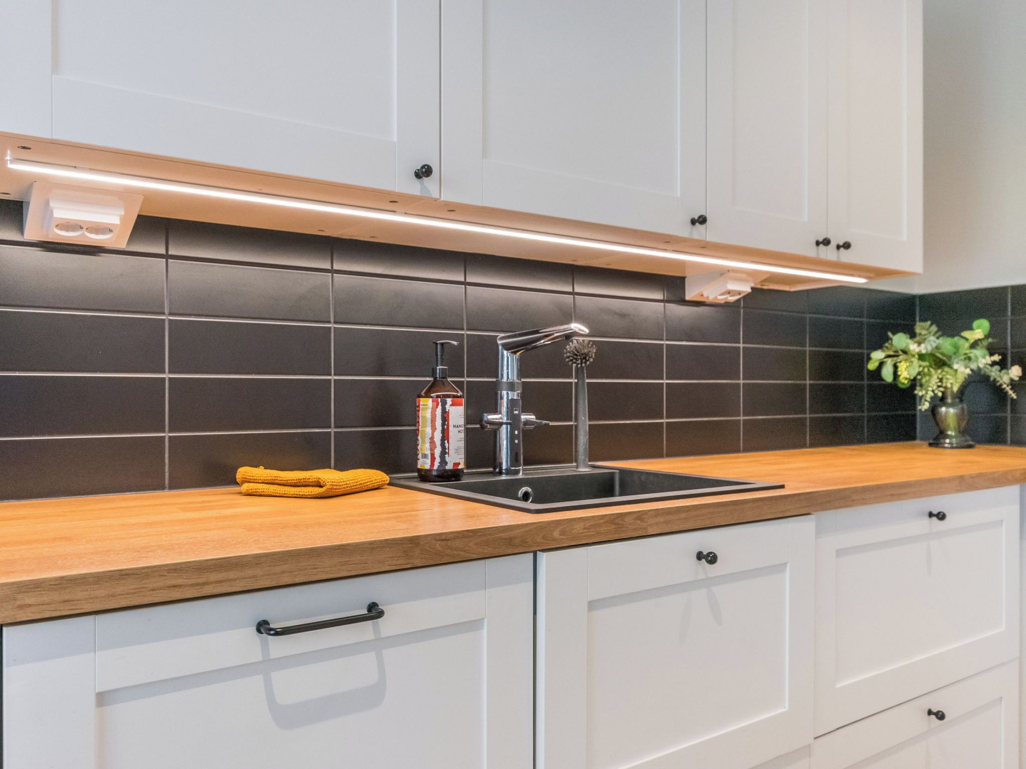 A modern kitchen with a stainless steel sink, wooden countertop, and black tiled backsplash.