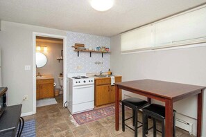Een kleine keuken met een fornuis, wastafel, houten kastjes en een eethoek met een tafel en krukjes.