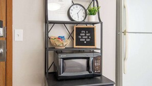 A kitchen area with a microwave on a black metal shelf, a clock, a potted plant, and a refrigerator. - Cozy Clean Affordable near NDSU and Downtown (Fargo)