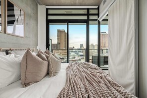Une chambre d’hôtel moderne dotée d’un grand lit, de plusieurs oreillers, d’une couverture en tricot et offrant une vue sur la ville grâce à de grandes fenêtres du sol au plafond.