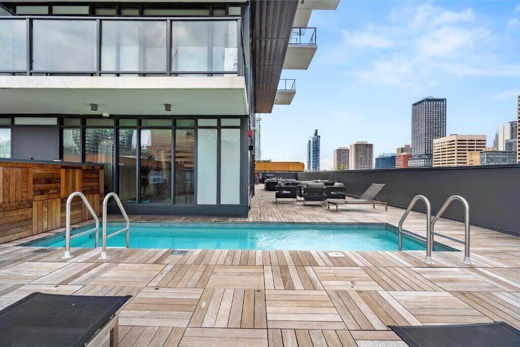 A rooftop pool area with a wooden deck, lounge chairs, and a city skyline in the background.