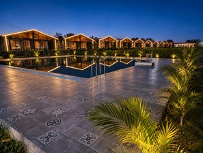A pool area with a tiled floor and a reflection pool, surrounded by multiple wooden buildings with glass windows. - Shreephal Luxurious Resort (Hatgad, Nashik)