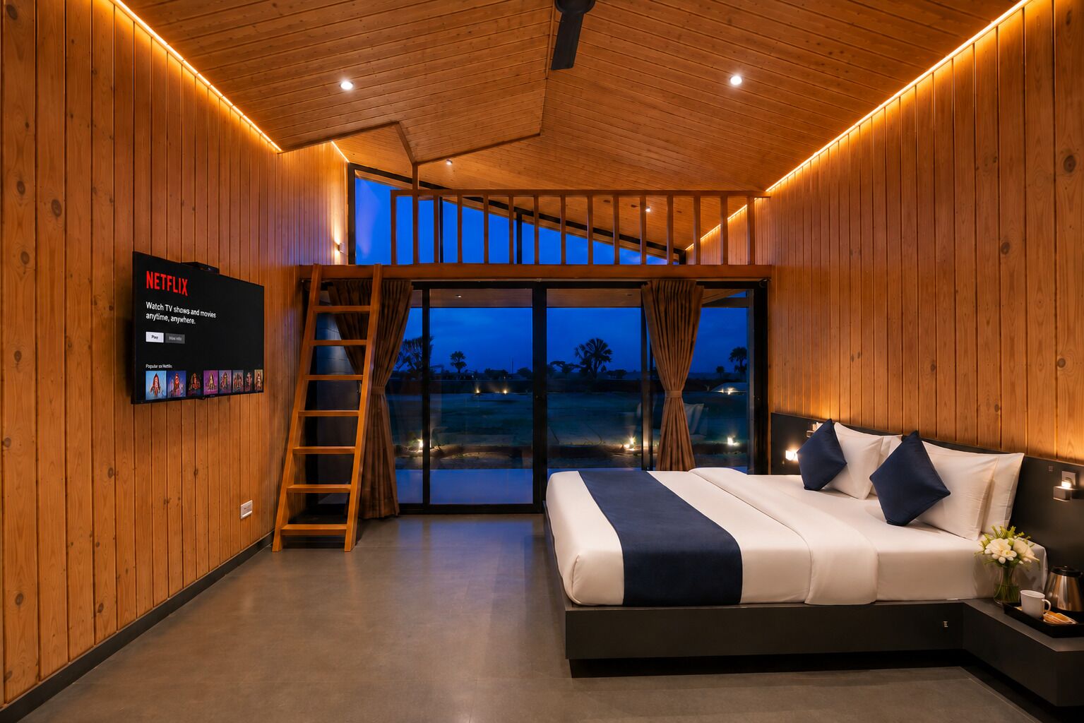 A modern bedroom with a large bed, a TV mounted on the wall, a sliding glass door leading to a balcony, and a wooden ceiling.