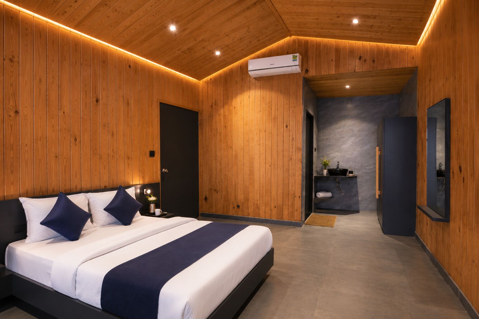 Un dormitorio moderno con techo de madera, una cama con ropa de cama blanca y azul marino, una mesita de noche y un baño con lavamanos y espejo.