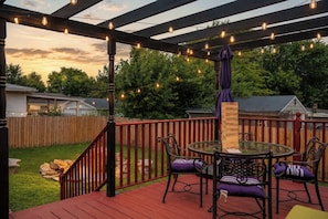 A deck with a glass table and chairs, surrounded by a wooden fence, under a pergola with string lights.