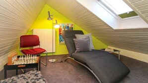 A small, triangular attic bedroom with a slanted ceiling, a red chair, a bed with two pillows, a chessboard, and a painting on the wall. - Townhouse Thaden - mid-terrace house in Norddeich for up to 4 people (Norden)