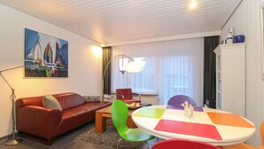 A modern living room with a brown leather sofa, a round table with colorful chairs, a floor lamp, and a painting on the wall. - Townhouse Thaden - mid-terrace house in Norddeich for up to 4 people (Norden)
